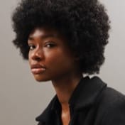 A person with a natural afro hairstyle wearing a black jacket looks at the camera against a plain background.